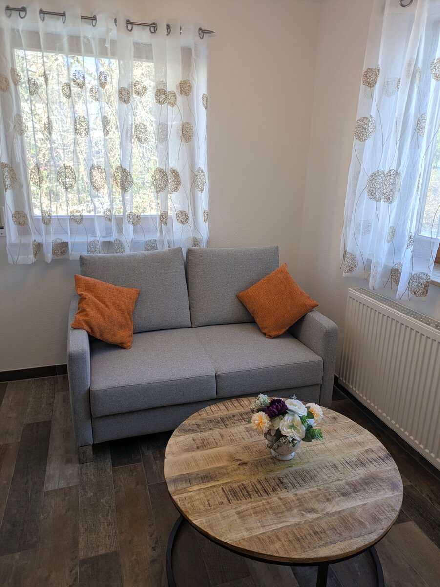 Graues Zweisitzer-Sofa mit orangefarbenen Kissen und rundem Holztisch in der Naturzeit Ferienwohnung