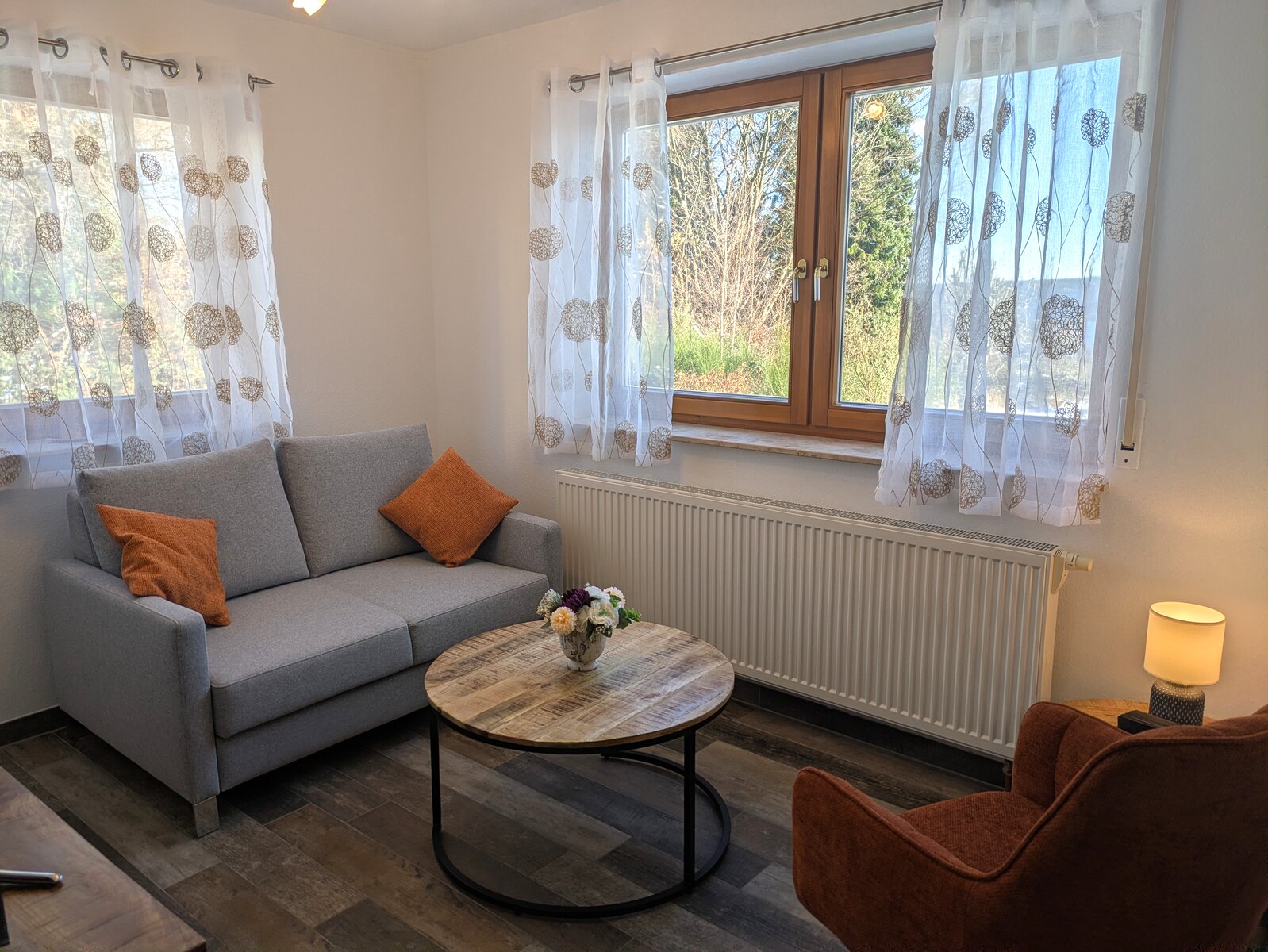 Gemütliches Wohnzimmer der Naturzeit Ferienwohnung mit grauem Sofa, orangefarbenen Kissen und Blick auf den Schwarzwald